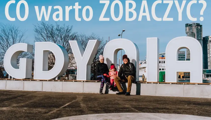 Rodzina pozuje przy napisie GDYNIA. W tle nowoczesne budynki, idealne na wakacyjne gdynia atrakcje dla dorosłych.
