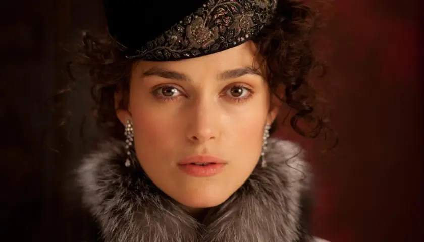 Anna Karenina w eleganckim futrze i ozdobnym kapeluszu, z błyszczącymi kolczykami, patrzy wprost przed siebie.
