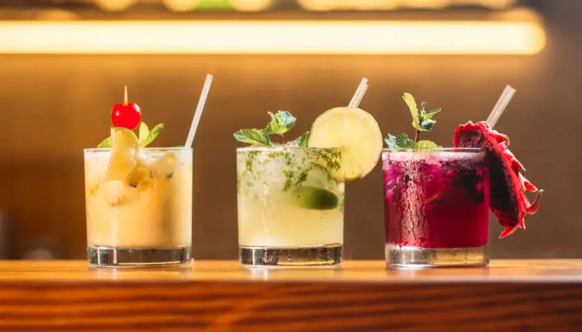 Trzy kolorowe drinki na barze: żółty z wiśnią, zielony z limonką i miętą, oraz fioletowy z pitają. Idealne do letnich wieczorów, a może i z **kazbek peak brandy z czym pić**?