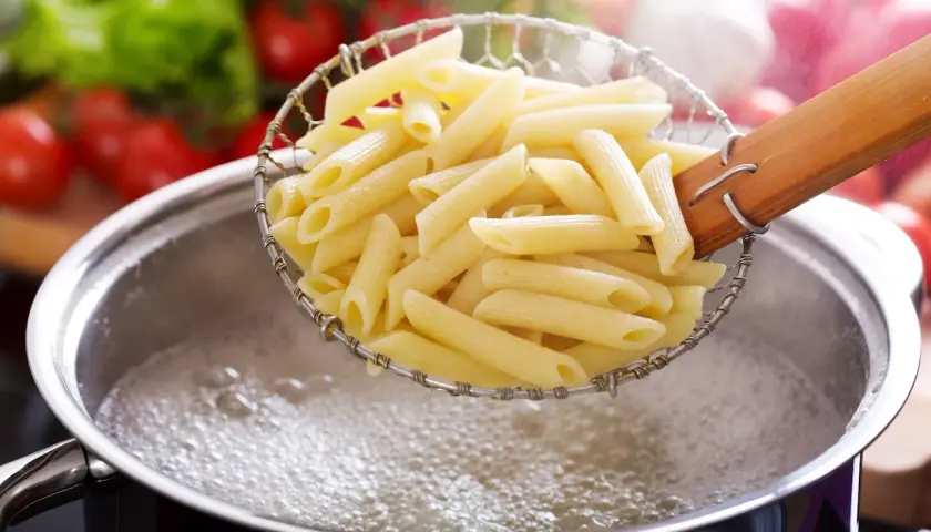 Gotowy do gotowania makaron penne w cedzaku nad bulgoczącym garnkiem. W tle pomidory i sałata.