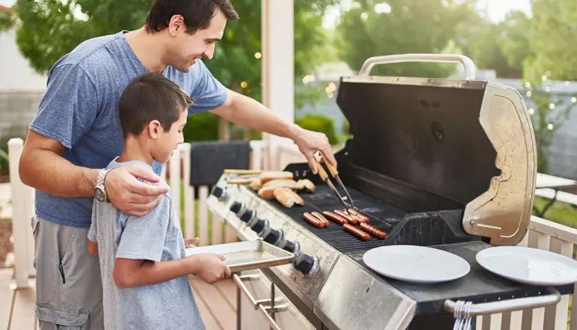 Un père et son fils préparent des saucisses sur un barbecue à gaz. Le père utilise des pinces pour retourner les saucisses pendant que le fils tient un plateau.