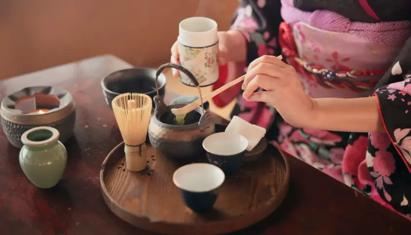 Eine Person in einem Kimono bereitet eine traditionelle matcha zeremonie vor. Ein Chasen (Bambusbesen) und eine Schale mit Matcha-Pulver sind auf einem Holzteller arrangiert.