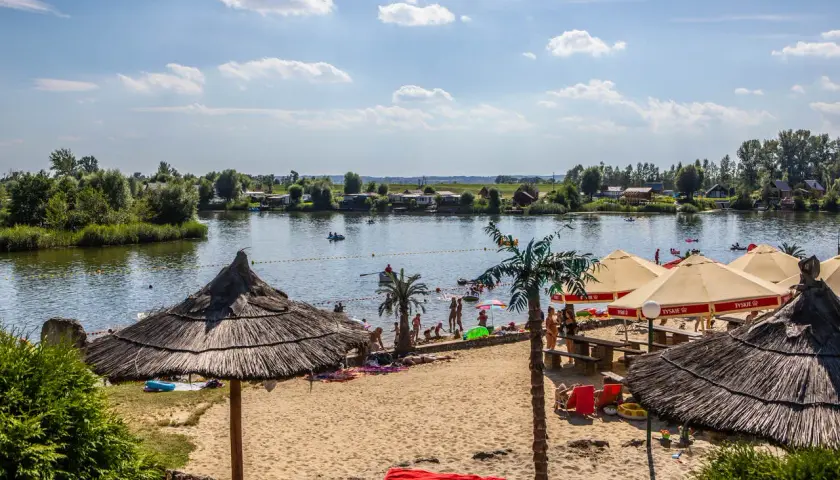 Słoneczny dzień na plaży w Kuźni Raciborskiej. Ludzie odpoczywają pod parasolami, kąpią się w jeziorze i pływają łódkami.