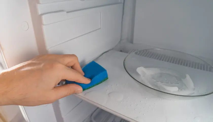 Mano limpiando el interior de una nevera con una esponja azul. El estante está mojado y hay un plato con hielo.