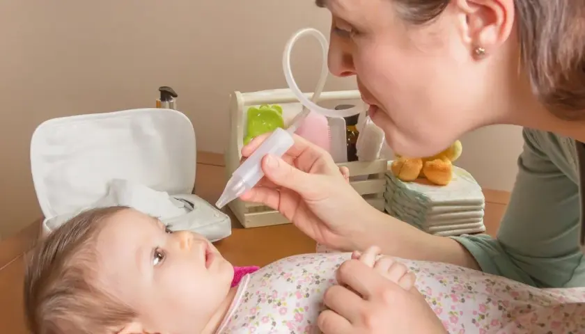 Mama pomaga noworodkowi z katarem, używając aspiratora. Dziecko leży na różowym ręczniku, patrząc na mamę.