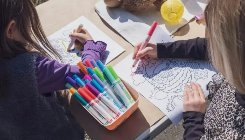 Zwei Kinder malen Osterbilder. Ein bunter Kasten mit Stiften liegt bereit, wie ein Laufzettel für die Grundschule.