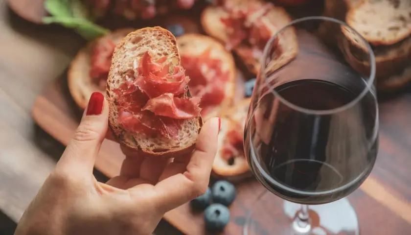 Une main tient une tartine garnie de jambon cru, à côté d'un verre de vin rouge. Parfait pour un apéro dinatoire.