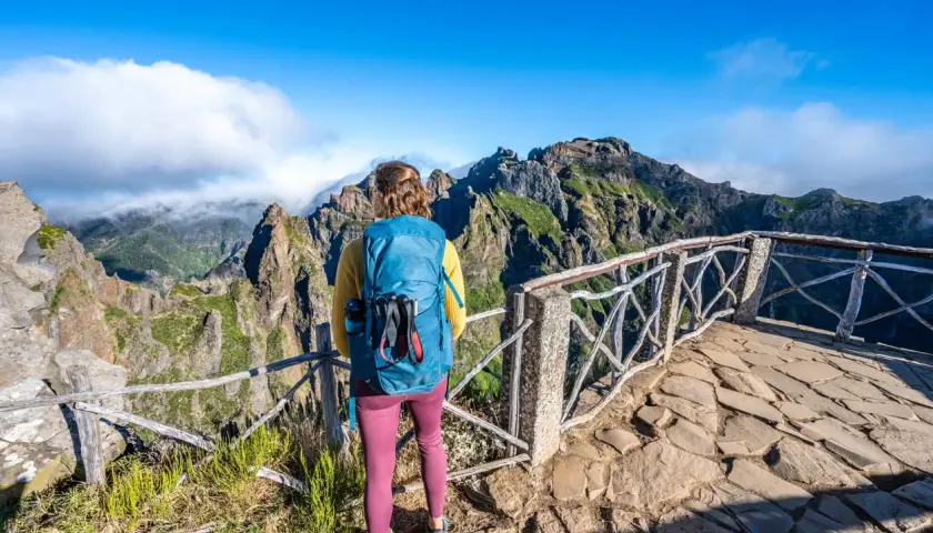 A hiker with a blue backpack enjoys a breathtaking view during a Madeira hike. Jagged, green mountains rise under a bright blue sky.