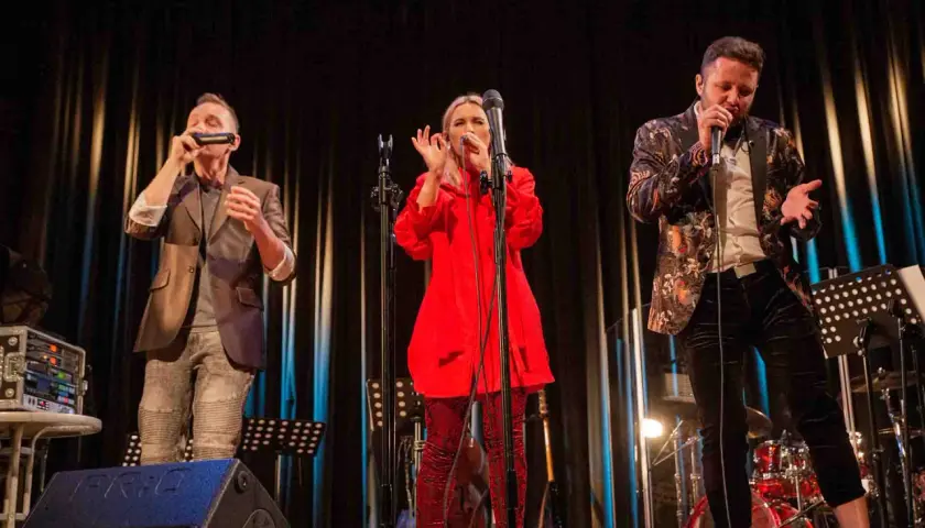 Trzech wokalistów na scenie podczas warsztatów wokalnych. Kobieta w czerwieni, mężczyzna w marynarce z wzorami i mężczyzna grający na harmonijce.