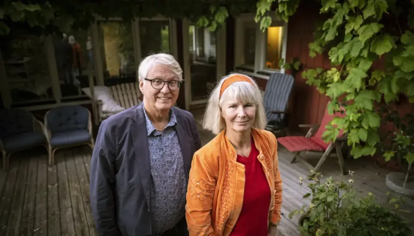 Ett par står på en altan. Mannen har glasögon och en blå kavaj. Kvinnan har ett orange hårband och en orange jacka. De ser ut som en del av amanda romare familj.