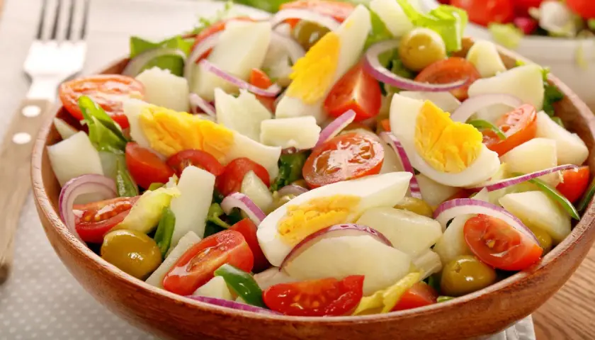 Deliciosa ensalada de patata con huevo duro, tomate, cebolla morada y aceitunas verdes en un cuenco de madera.