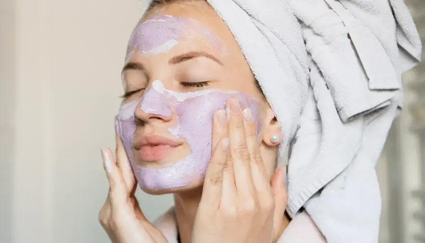 En kvinna med handduk i håret applicerar en lila ansiktsmask. En avkopplande ansiktsbehandling hemma för att skämma bort sig själv.