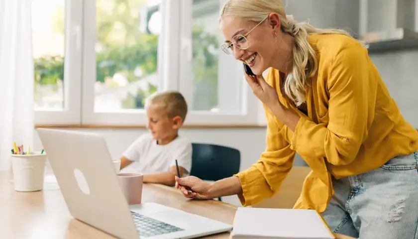 Uśmiechnięta kobieta rozmawia przez telefon, pracując przy laptopie, podczas gdy jej syn rysuje obok. Odpowiedź na pytanie: czy na urlopie wychowawczym można pracować.