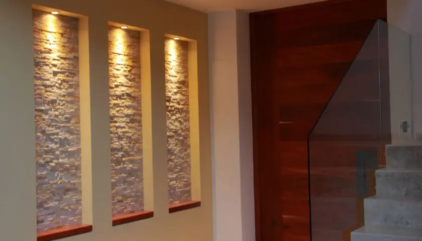 Tre nicchie in cartongesso illuminate da faretti, con rivestimento in pietra, creano un punto focale moderno accanto a una porta in legno e una scala con parapetto in vetro.