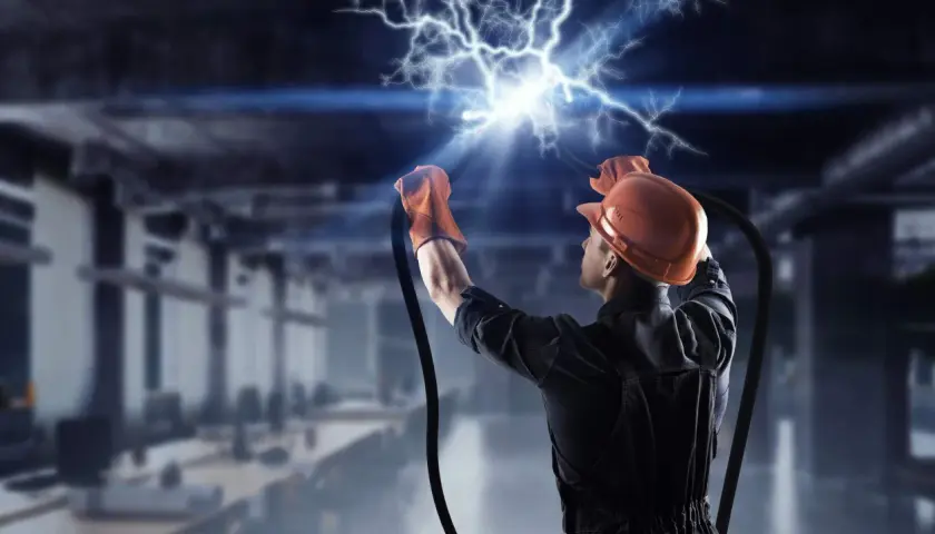 Elektryk w kasku i rękawicach trzyma przewody, z których wydobywa się potężne przepięcie elektryczne.