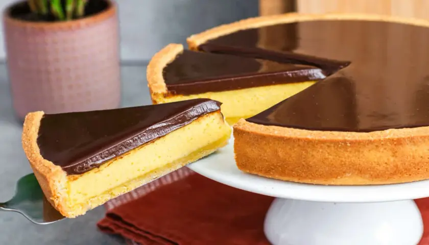 Una fetta di torta Susanna, con crema gialla e copertura al cioccolato fondente, viene sollevata da una tortiera su un'alzata bianca.