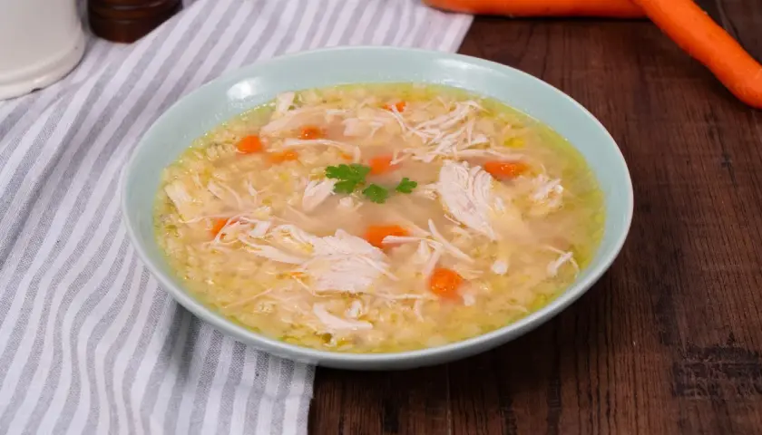 Un confortante brodo di pollo con pezzi di pollo sfilacciato, carote e un rametto di prezzemolo.