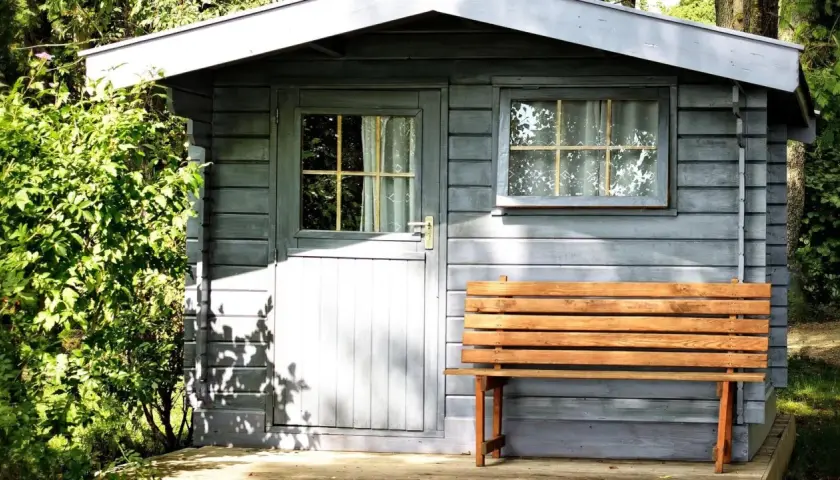 Ein kleines Gartenhaus mit Holzbänken davor. Das Dach muss dringend abgedichtet werden, um es vor Regen zu schützen.