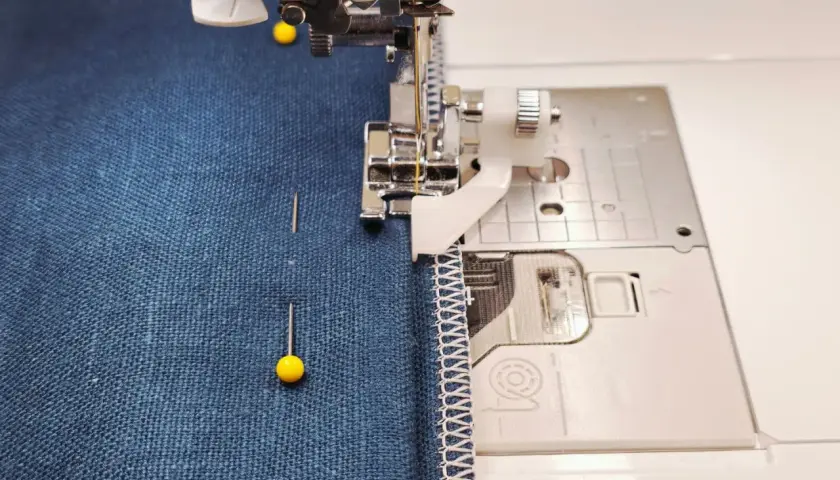 A sewing machine with a blind stitch foot is about to hem dark blue fabric. Pins hold the fabric in place.