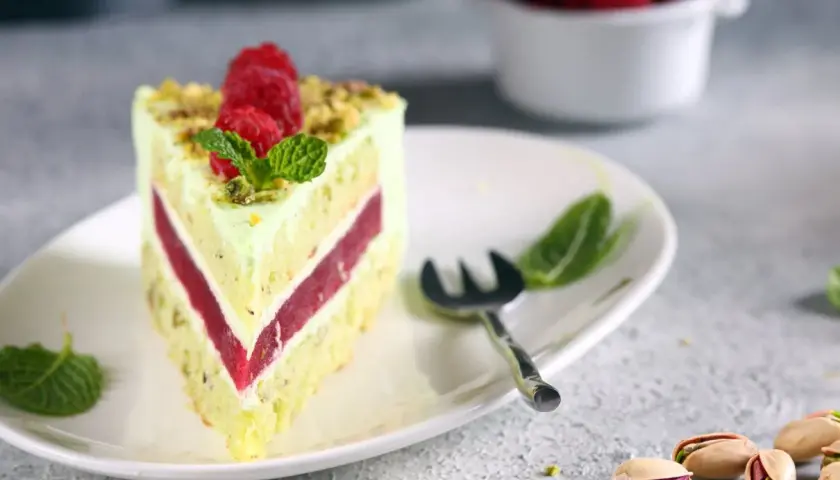 Pyszny tort pistacjowo malinowy z malinami i miętą na talerzu. Obok widoczne są pistacje.