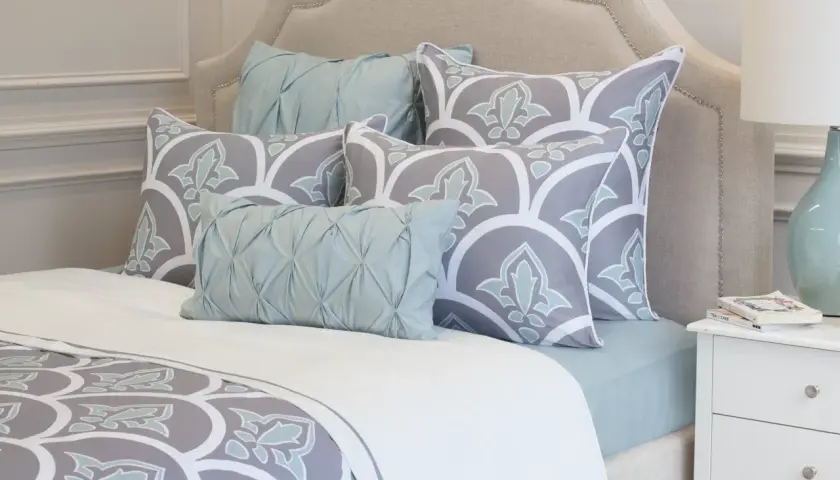 A bed with a tufted headboard, featuring large square pillows with a grey and white floral pattern, and smaller light blue accent pillows.