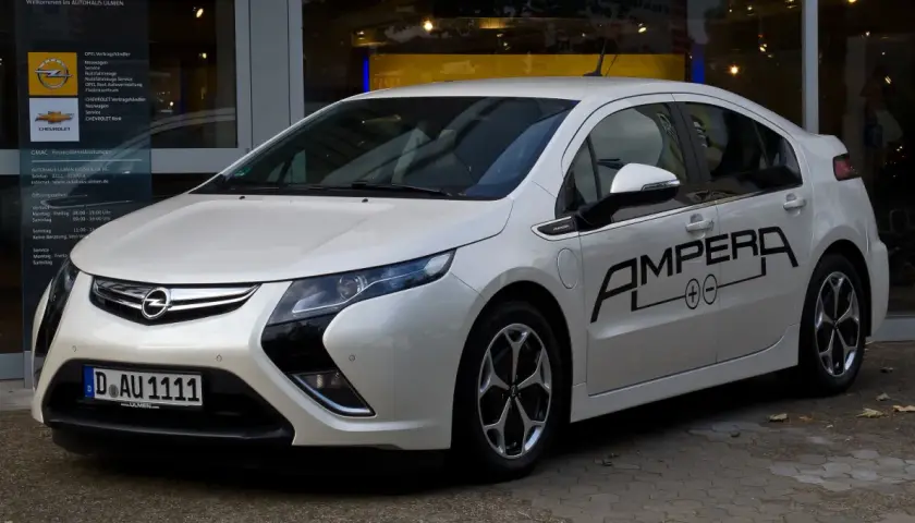 Biała Opel Ampera z czarnym napisem "AMPERA" na drzwiach, zaparkowana przed salonem samochodowym.