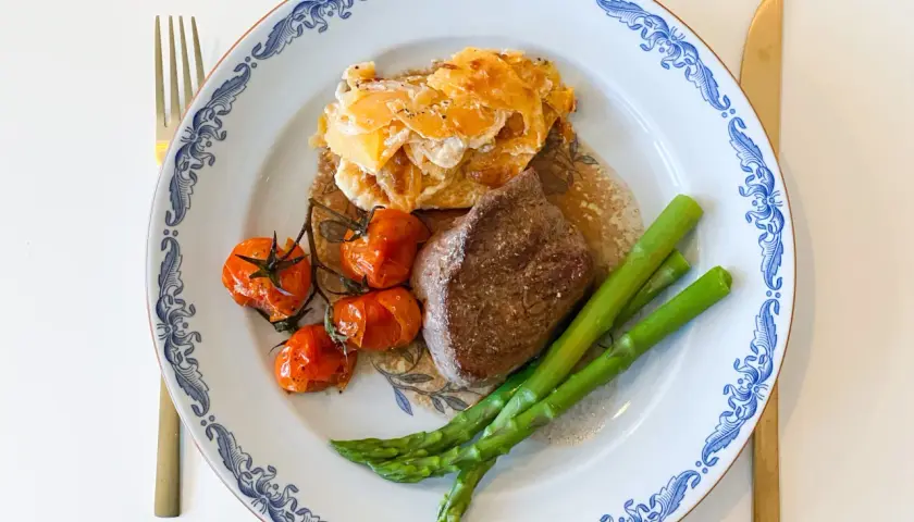 En tallrik med oxfilé sous vide, gratinerad potatis, körsbärstomater och sparris. Guldfärgad kniv och gaffel.