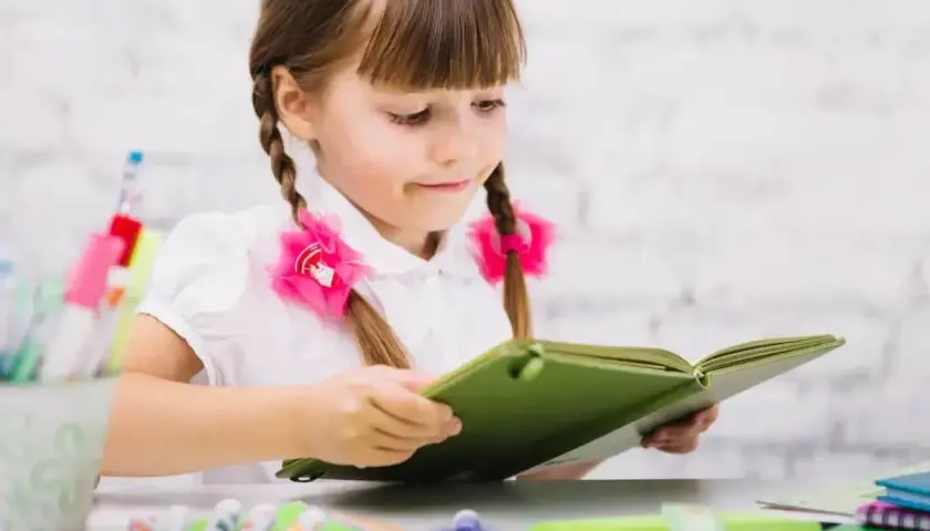 Dziewczynka z warkoczami i różowymi kokardkami pochłonięta lekturą. Jej przygotowanie pedagogiczne widać w skupieniu na nauce.