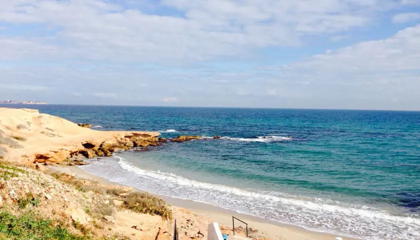 Escaleras descienden a la playa rocamar, con arena dorada, rocas y mar azul bajo un cielo nublado.