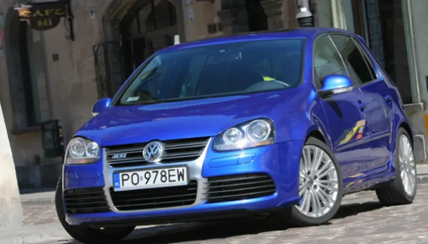 Błękitny Golf R32 z felgami ze stopów lekkich, zaparkowany na brukowanej uliczce przed kawiarnią.