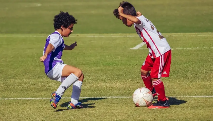 Due ragazzini si sfidano su un campo da calcio in erba, uno in maglia viola, l'altro in maglia bianca e rossa, con un pallone ai loro piedi.
