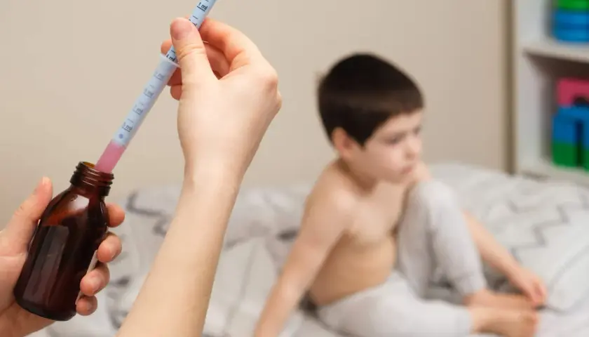 Mano preparando jeringa con ibuprofeno para un bebé. Niño en cama al fondo.