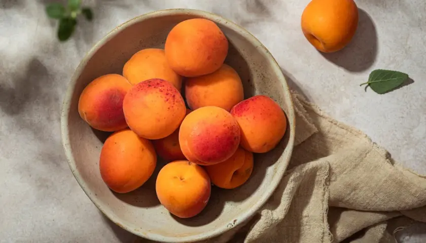 Un bol rempli d'abricots juteux, annonçant la belle **période abricot**. Un fruit isolé repose à côté, sur un tissu beige.