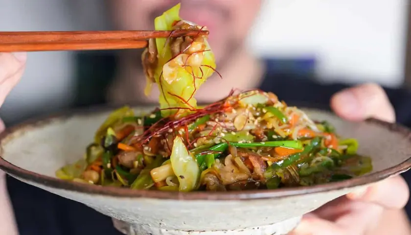 A person uses chopsticks to lift a bite of a colorful Japanese stir fry, featuring cabbage, carrots, and peppers, from a rustic bowl.
