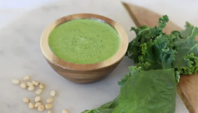 Un cuenco de madera contiene un vibrante pesto de kale, acompañado de hojas frescas de kale, albahaca y piñones.