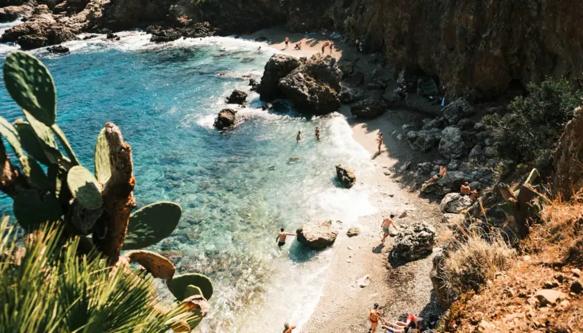 A secluded beach in Zingaro, Sicily, with crystal-clear turquoise water, rocky cliffs, and sunbathers enjoying the Mediterranean.