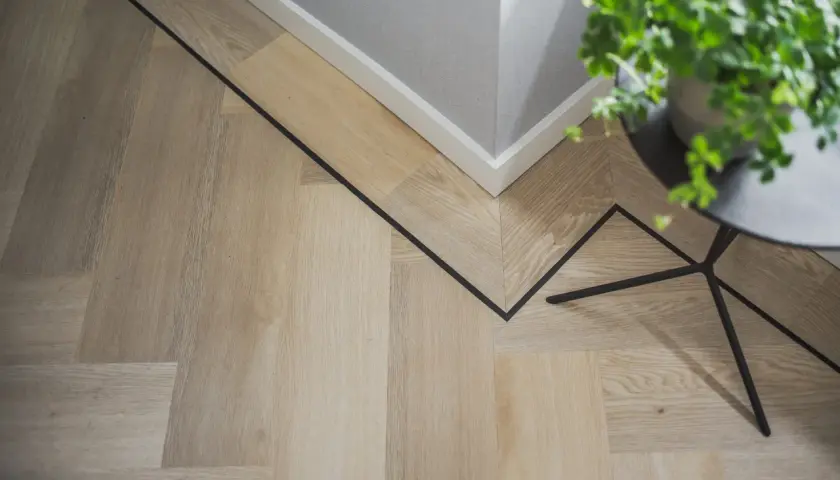 Drewniana podłoga w jodełkę z czarną, geometryczną wstawką. W rogu stoi doniczka z zieloną rośliną, a obok czarny stolik.