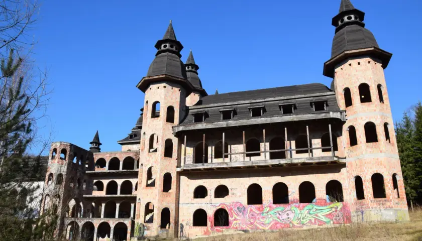 Niedokończony zamek w Łapalicach, z ceglanymi wieżami i łukowymi oknami, pokryty graffiti, pod błękitnym niebem.