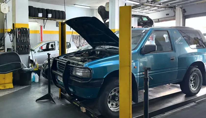 Taller mecánico con coches, neumáticos y herramientas. Se ven las copelas de un coche azul, parte del motor y la carrocería.