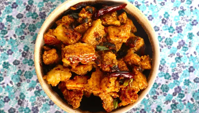 Crispy, golden-brown pieces of ice tofu are coated in a savory sauce with chili peppers and green onions, served in a bowl on a floral background.