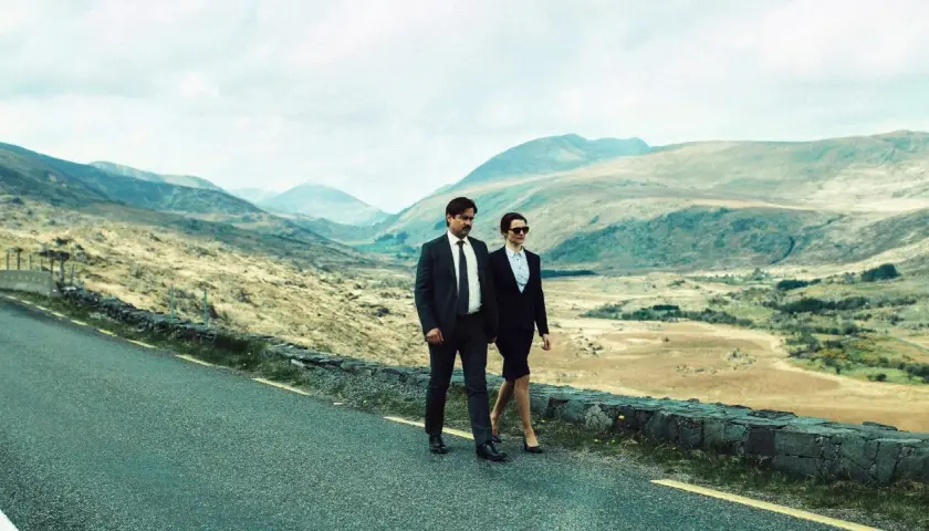 Coppia in abiti eleganti cammina su strada panoramica, evocando il significato di "The Lobster" e la ricerca di un legame in un paesaggio desolato.