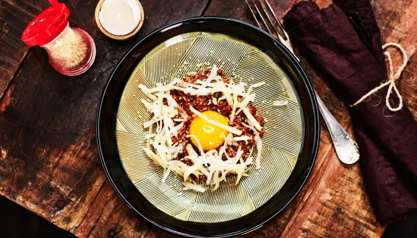 En läcker koreansk råbiff med en gul äggula i mitten, toppad med strimlad rättika och sesamfrön. Serveras på en vacker tallrik med en mörk servett och gaffel.