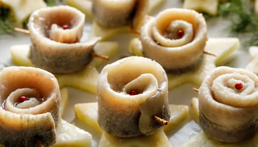 Roladki ze śledzi w occie, ozdobione ziarnami pieprzu, ułożone na plasterkach jabłka.