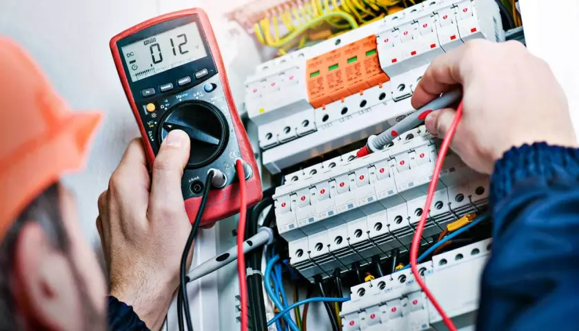 Elektryk w pomarańczowym kasku sprawdza instalację multimetrem. To pierwszy krok, jak zostać elektrykiem.