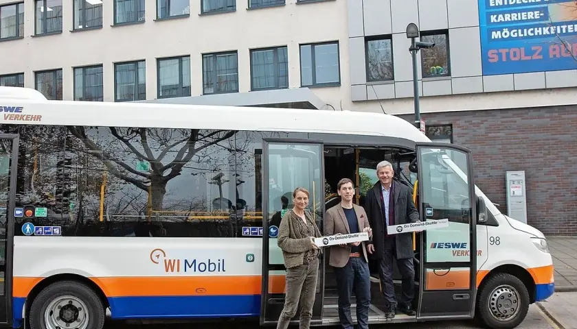 Drei Personen stehen vor einem weißen Kleinbus mit der Aufschrift "WI Mobil". Sie halten Schilder mit der Aufschrift "On-Demand-Shuttle".