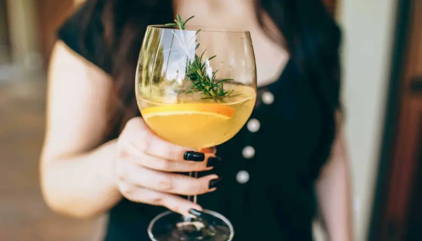 En person håller ett glas med en uppfriskande drink med vitt vin, garnerad med en apelsinskiva och en rosmarinkvist.
