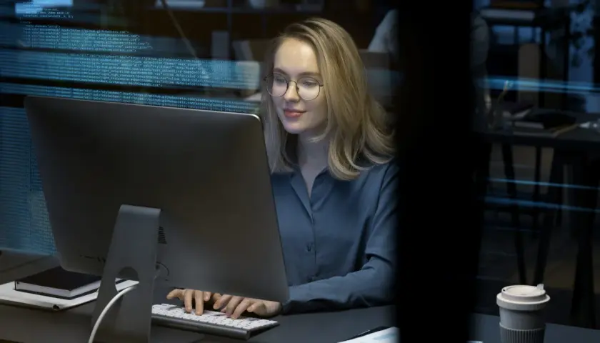 Młoda kobieta w okularach pracuje przy komputerze, na ekranie widać kod. To przyszłość cyberbezpieczeństwa, studia w tej dziedzinie otwierają wiele możliwości.
