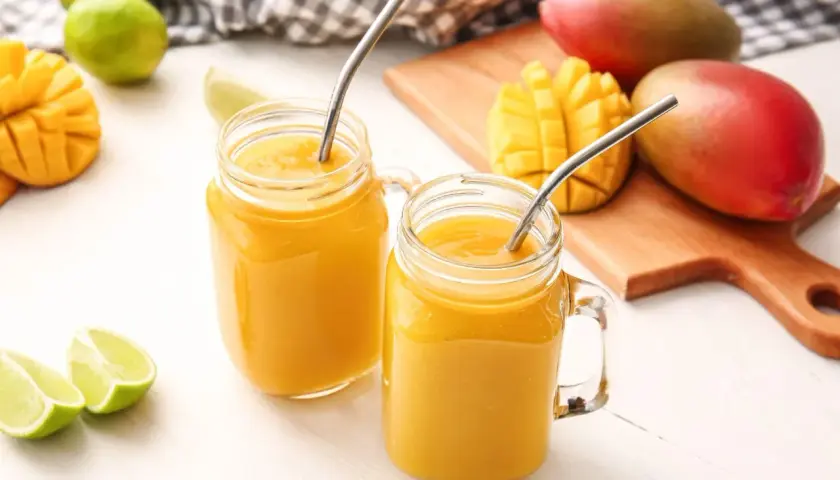Dwa orzeźwiające drinki z mango w słoikach z metalowymi słomkami, obok świeżych mango i limonek.