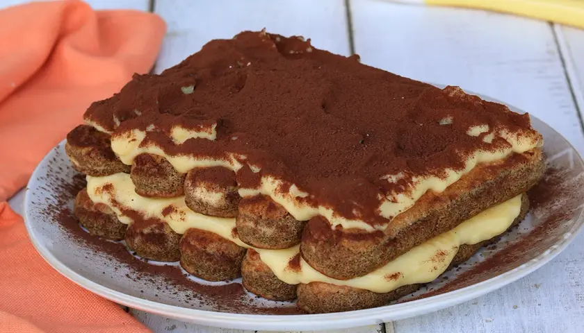 Tiramisù con crema pasticcera, strati di savoiardi e cacao in polvere su un piatto bianco, con un tovagliolo arancione sullo sfondo.