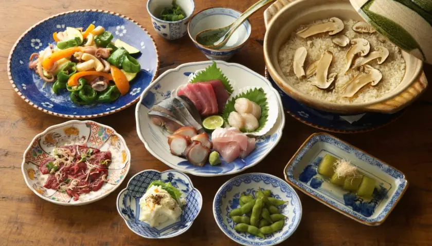 A spread of Japanese side dishes, including sashimi, edamame, potato salad, and a vegetable stir-fry, complements a pot of rice with mushrooms.
